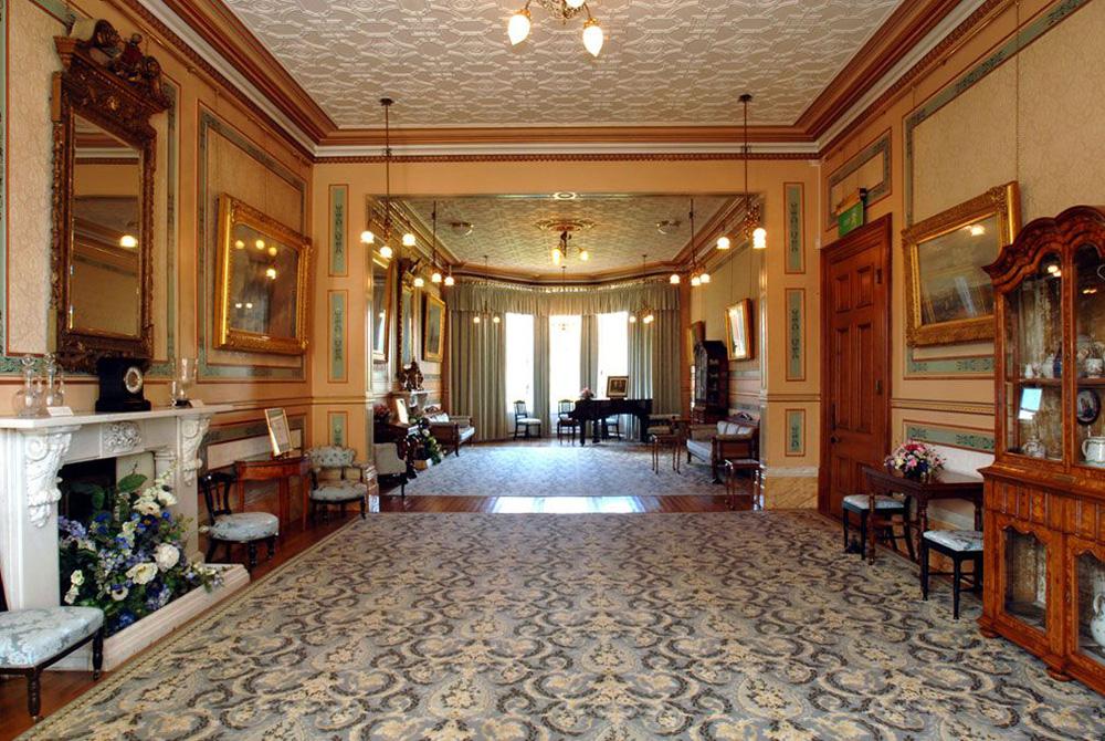 A ceremony room with a patterned carpet, fireplace, mirror, grand piano, framed artwork on the walls, and several chairs and tables and large curtained windows.
