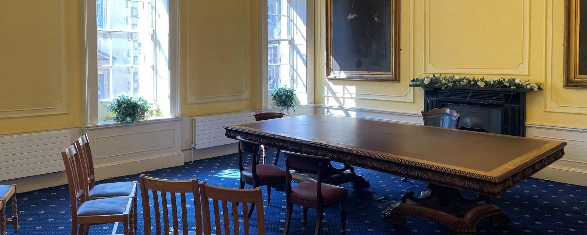 A ceremony room with yellow walls, large wooden table, chairs, tall windows overlooking St Nicholas Market, blue carpet, potted plants and a decorative fireplace.