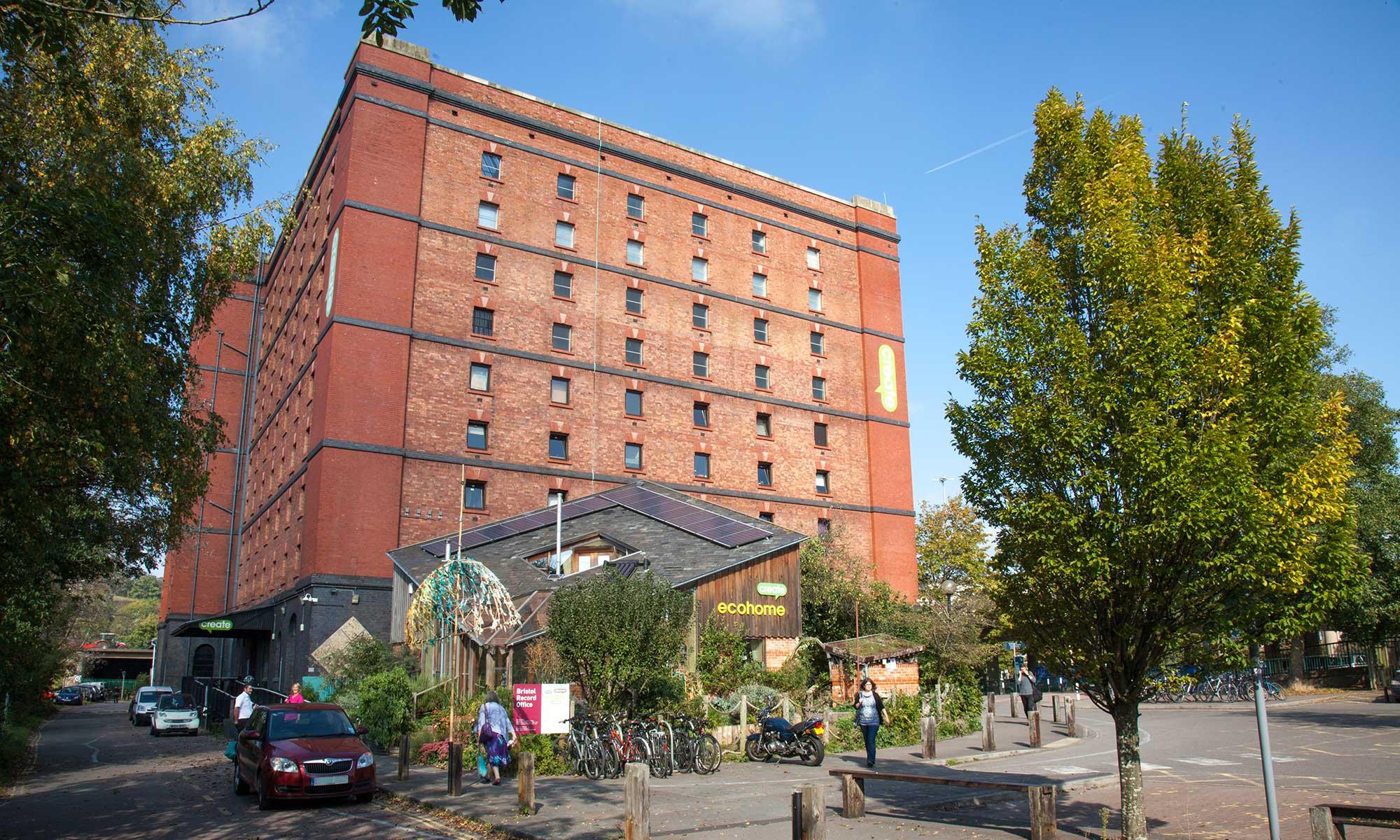 Large red‑brick multi‑storey building with rows of small windows. Trees, bicycles, parked cars, and pedestrians in the foreground